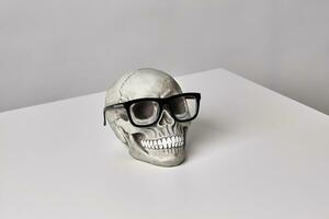 Realistic model of a human skull with teeth in a black-framed classic glasses on a light table, white background. Halloween horror concept. photo