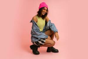 Fashion studio photo of black smiling woman in jeans jacket and pink hat  posing on pink background.