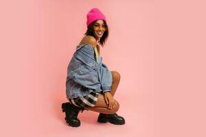 Fashion studio photo of black smiling woman in jeans jacket and pink hat  posing on pink background.