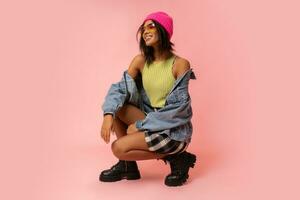 Fashion studio photo of black smiling woman in jeans jacket and pink hat  posing on pink background.