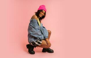 Fashion studio photo of black smiling woman in jeans jacket and pink hat  posing on pink background.