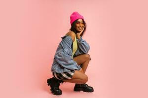 Fashion studio photo of black smiling woman in jeans jacket and pink hat  posing on pink background.