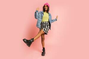 Stylish  fancy african woman in pink hat , cool sunglasses and  jeans jacket posing on pink background. Winter fashion look. Full lenght. photo