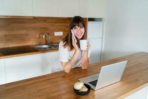 sonriente europeo mujer hablando por móvil teléfono y Bebiendo café en moderno cocina. foto