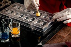 A woman confectioner with red uniform and white sterile gloves do a set of colorful chocolates from milk chocolate on a table. photo