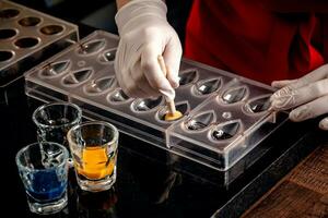 A woman confectioner with red uniform and white sterile gloves do a set of colorful chocolates from milk chocolate on a table. photo