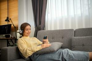 asiático mujer sentar en el sofá en el vivo habitación a hogar a disfrutar su favorito canciones niña usa auriculares a escucha a música, estado animico y pasatiempo, moderno inalámbrico tecnología concepto foto