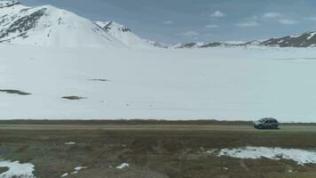 SUV Car Goes on High Altitude Gravel Road in Snowy Mountains at Sunny Day. Aerial View. Drone is Flying Sideways and Distancing. Establishing Shot video