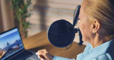 A blonde woman in a blue shirt hosts a podcast from her home studio. There is a computer and notepads on the table. Lively communication video