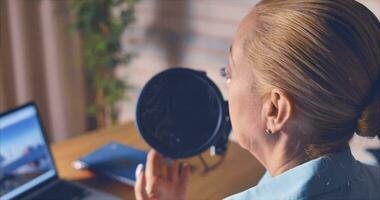 A woman in a blue shirt hosts a podcast from her home studio. There is a computer and notepads on the table. Lively communication video