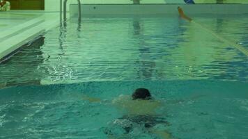 atlético caucásico hombre con gorra y gafas de protección es nadando braza de pecho adelante en piscina con azul agua. espalda vista. video