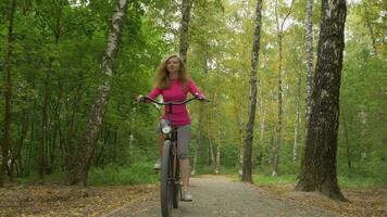 Jeune souriant femme est équitation vélo dans le parc avec déchue feuilles dans l'automne vers le caméra. steadicam tir. faible angle. lent mouvement. video