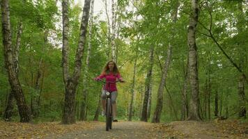 Young woman is riding bicycle in the park with fallen leaves in autumn towards the camera. Steadicam shot. Low angle. video