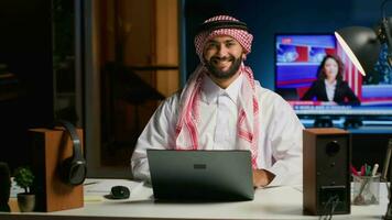 retrato do sorridente árabe homem de negocios trabalhando às moderno mesa, digitando em dele computador portátil, resolução tarefas. muçulmano cara navegando em digital dispositivo, fazendo o email comunicação dentro profissional escritório configuração video