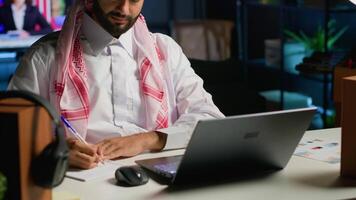 árabe empreendedor trabalhando às casa escritório escrivaninha em computador portátil, navegando a Internet e escrevendo dentro caderno. muçulmano homem usando caneta, bloco de anotações e digital dispositivo enquanto teletrabalho video