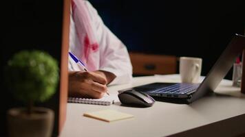 Man in apartment paying attention in elearning seminar, writing on notepad using pen. Middle Eastern homeschooling student listening to lesson while doing his homework tasks, close up video