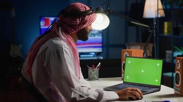 Arab man browsing on mockup laptop while working from home. Self employed freelancer checking emails on isolated screen device in apartment office with TV as background noise video