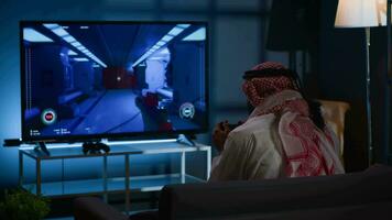 arabe homme en jouant jeux vidéos sur la télé, relaxant après longue journée à travail. compétitif joueur ayant amusement sur jeu console dans élégant appartement vivant chambre, profiter science fiction tireur Jeu video