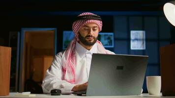 árabe empregado concentrando em acabamento acima importante trabalho tarefas, remotamente trabalhando a partir de à moda apartamento pessoal escritório. focado muçulmano teletrabalhador digitando em computador portátil teclado video