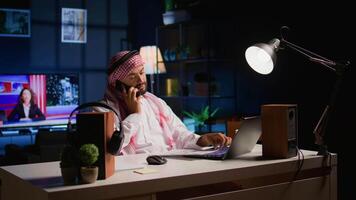 Arab worker answering phone call while remotely working from cozy home office. Teleworker talking with colleague using smartphone, discussing paperwork and tasks needing to be solved video