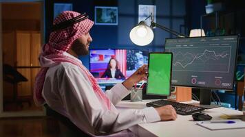 Muslim remote worker at home holds chroma key tablet while looking at business annual report graphs on computer. Arabic freelancer with mockup digital device in hand performing company tasks video