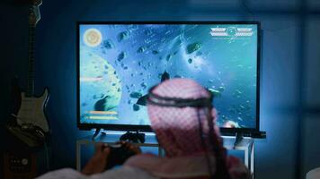 Middle Eastern gamer plays classic arcade action videogame, flying through space debris. Man enjoying leisure time in living room using high tech gaming console to solve missions in singleplayer game video