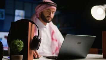 Arabic teleworker remotely working in dimly lit apartment sending emails on laptop. Middle Eastern worker at home typing business data on notebook device, wearing traditional attire video