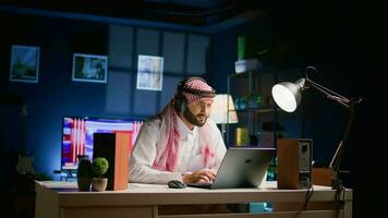 Arab man with wireless headphones at home in living room, watching educational videos on laptop. Middle Eastern person listening to podcasts, enjoying relaxing leisure time using notebook