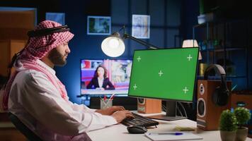 Arabic man looking at chroma key computer screen while working from home. Self employed person typing on keyboard, sending emails on green screen device in apartment office with TV as background noise video