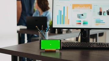 Empty workstation with greenscreen display on mobile phone app, desktop showing marketing analysis and reports in coworking space. Business office desk displaying smartphone screen with chromakey. video