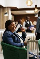 Happy smiling African American woman arriving at resort, sitting in hotel lobby with smartphone and waiting until checkin time. Black female guest sitting in lounge area using phone, traveling alone photo
