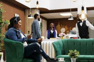 Happy young African American woman in headphones sitting relaxing in hotel lobby browsing internet on phone. Smiling female tourist listening music on smartphone while resting at resort photo