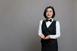 Smiling asian professional waitress showing with hands while looking at camera. Young attractive restaurant receptionist wearing black and white uniform suit and posing for studio portrait photo