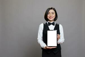 Restaurant asian woman receptionist holding digital tablet with white empty screen for catering service app. Smiling cheerful waitress in unform showing blank touchscreen for promotion photo