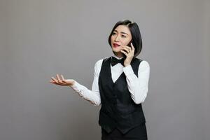 asiático mujer recepcionista chateando con cliente en teléfono inteligente y haciendo gestos con mano. joven asiático camarera vestido en negro y blanco uniforme teniendo comunicación en móvil teléfono foto