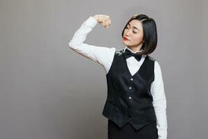 Proud asian waitress wearing professional uniform showing strong bicep muscle. Young attractive woman receptionist showcasing strength and power while posing in studio on gray background photo