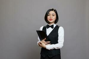 Confident young asian woman waitress wearing uniform and bow tie standing with digital tablet portrait. Serious receptionist holding portable gadget in hands and looking at camera photo