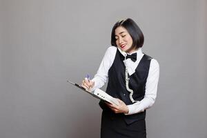 sonriente asiático camarera respondiendo teléfono fijo teléfono llamada y escritura en portapapeles. restaurante simpático mujer empleado en uniforme hablando con cliente en teléfono y gerente orden foto