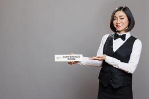 Smiling asian receptionist showing conference room direction sign and looking at camera. Hospitality service cheerful woman worker holding boardroom pointer sign studio portrait photo