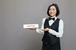 Restaurant receptionist showcasing reception steel plate, smiling and looking at camera with happy expression. Hotel asian woman worker showing tabletop sign studio portrait photo