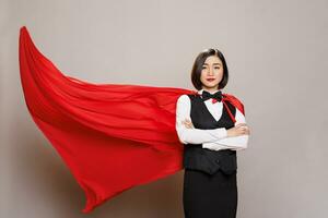 recepcionista con brazos cruzado vistiendo rojo superhombre capa, representando poder y confianza retrato. atractivo asiático camarera vestido en restaurante uniforme y revoloteando superhéroe capa foto