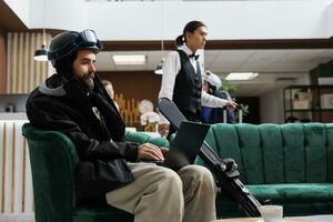 Male vacationer dressed in snow clothing sits on sofa holding his digital minicomputer in hotel reception. Caucasian man with skiing gear using laptop for online booking at ski mountain resort. photo