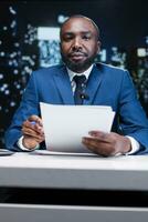 TV host reporting daily events news in studio, going live on midnight broadcast to discuss important incidents worldwide. African american journalist presenting information and details. photo