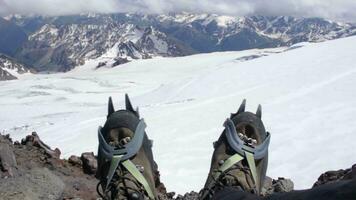 alpinista en crampones es sentado en un rotura y sacudida su piernas. Nevado montañas en antecedentes. primero punto vista. video