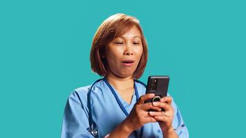 Close up of cheerful clinic employee texting on phone while at work, enjoying break. Upbeat BIPOC nurse wearing professional uniform chatting with friends online, isolated over studio background photo