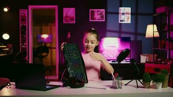 Child influencer filming keyboard, mouse and headphones review in home studio, showing capabilities and specifications. Little girl showcasing computer peripherals to fans on gen Z online channel video