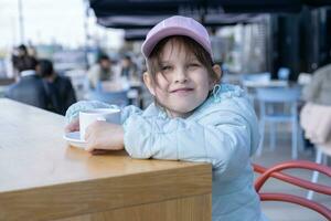 divertido linda niña en calle cafetería. pequeño niña Bebiendo caliente bebida foto