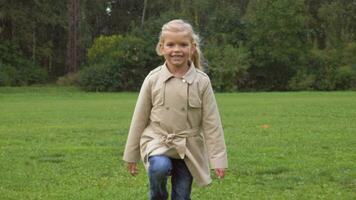 Small preschooler smiling caucasian girl in jacket and blue jeans is running on green lawn in the park towards the camera. Slow motion. video