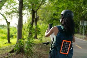 Rear view of young woman with helmet taking selfie with mobile phone in the park. It has a backpack with a solar panel for charging smartphone. photo