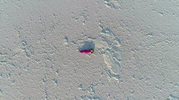 Young caucasian woman in red dress with long hairs lies on white surface of salt lake. Drone is fast flying upward. Aerial vertical shot.  . video